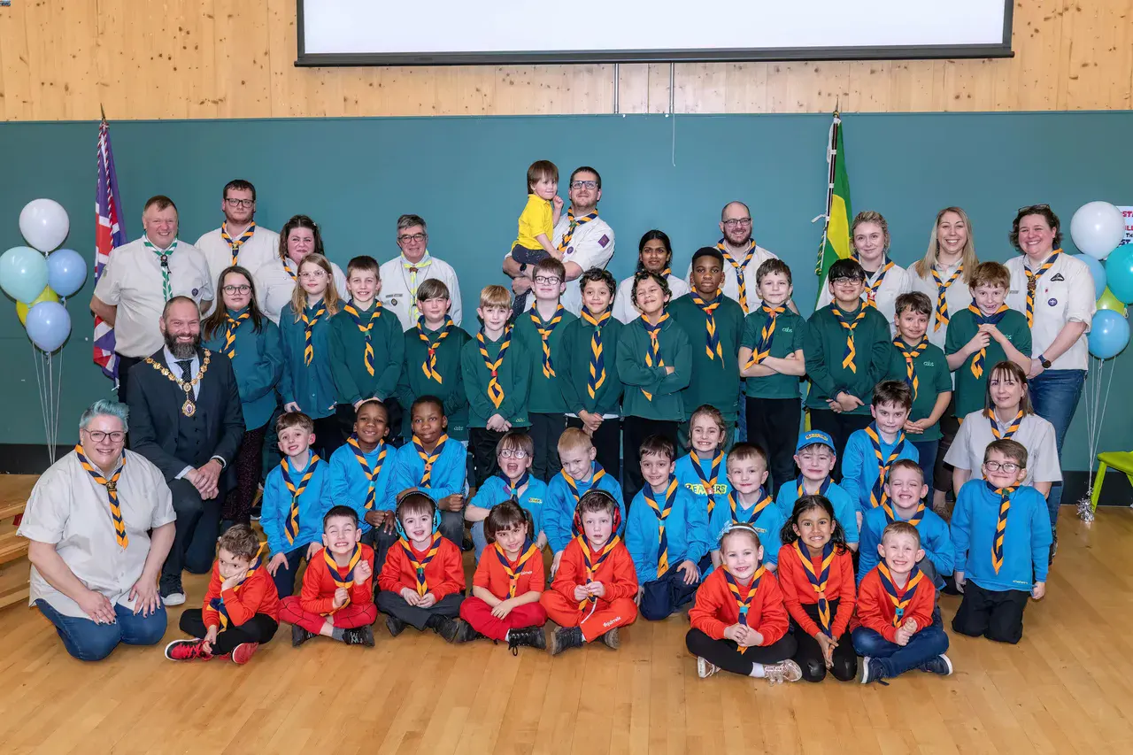A diverse group of scouts and leaders in bright uniforms pose smiling in a hall. Balloons and flags decorate the space, creating a cheerful atmosphere.