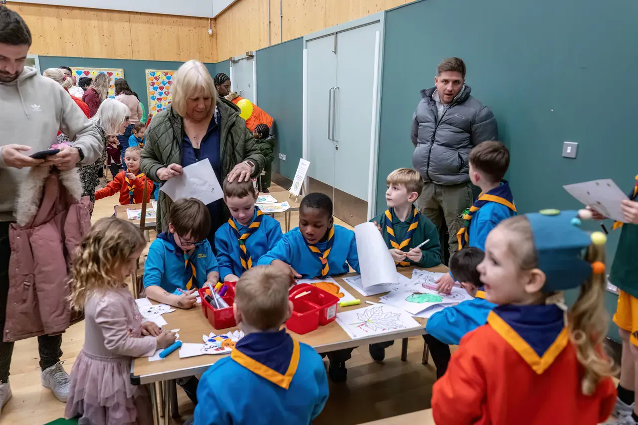 Children in blue uniforms with yellow scarves sit at tables doing arts and crafts in a classroom. Adults stand nearby, and colorful drawings are on the tables.