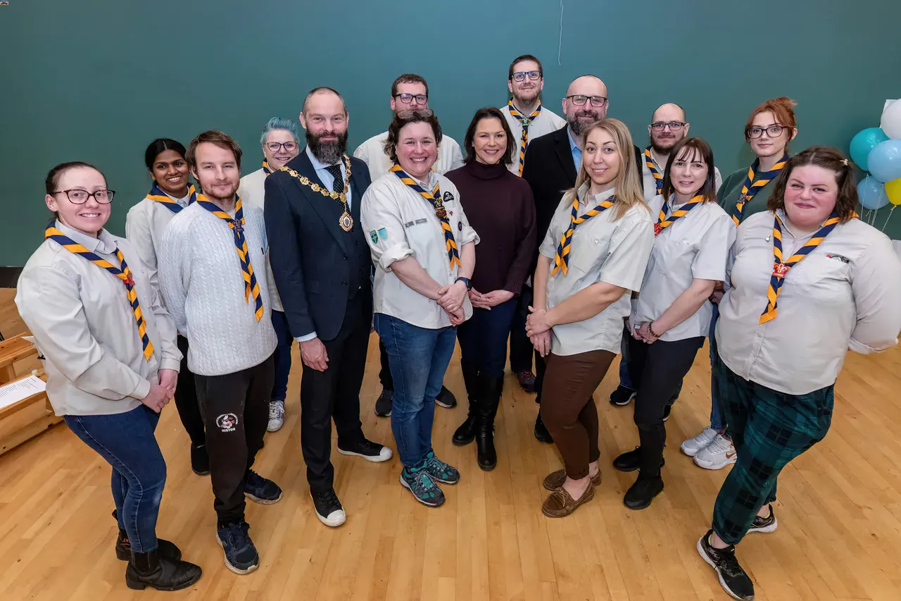 A group of people wearing scout uniforms with yellow and blue neckerchiefs stand smiling in a hall. A person in a suit with a chain is among them.