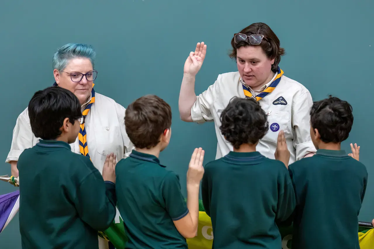 Two scout leaders, wearing uniforms and scarves, lead a ceremony with four children raising their right hands. The mood is formal and engaging.