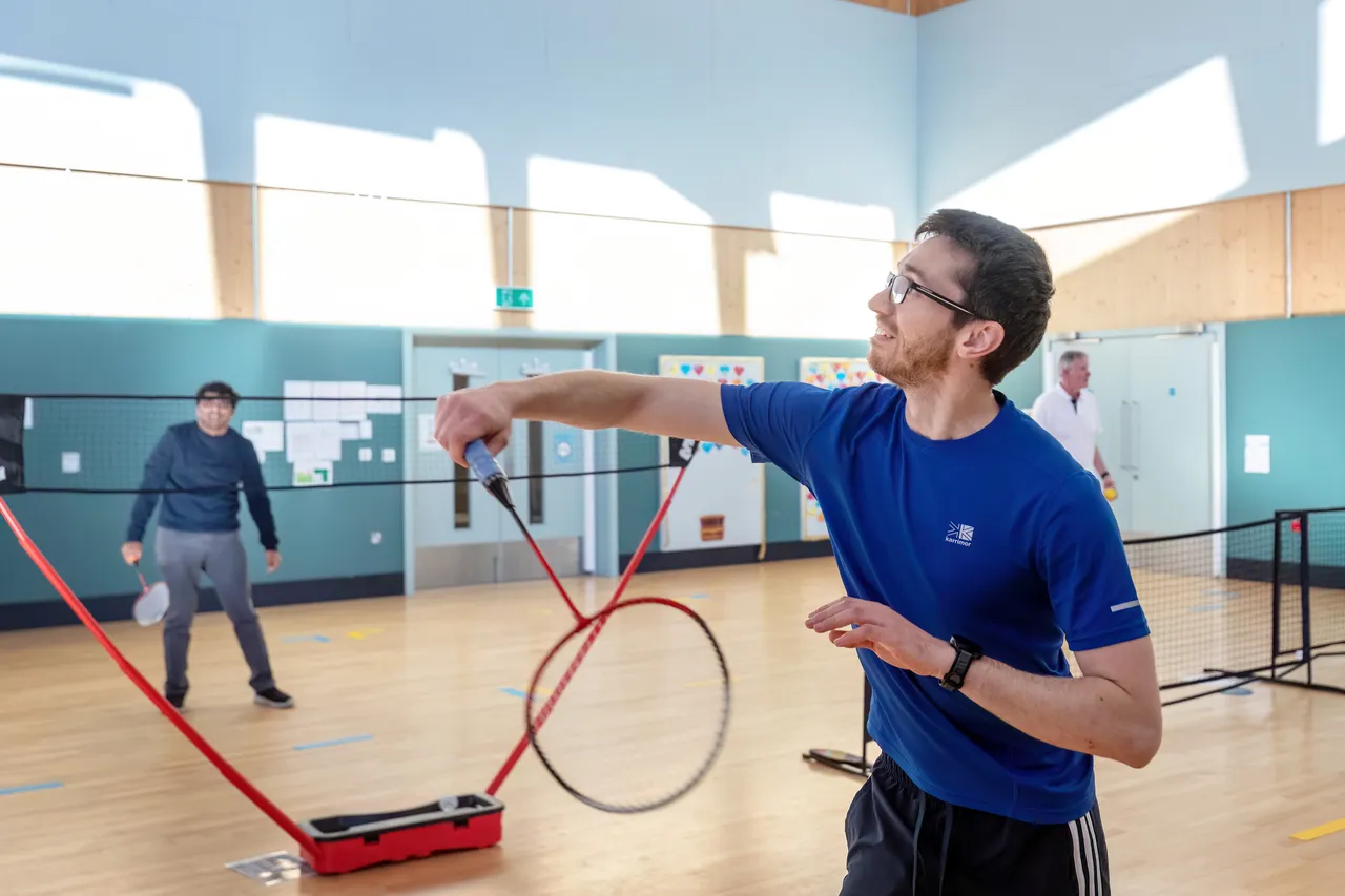 A man with glasses and a blue t-shirt smiles as he prepares to hit a shuttlecock with a red badminton racket. In the background, another man on a court prepares to return the shot across a portable black net. The setting is a bright, indoor sports hall with light wood floors and blue walls. The image represents Wintringham residents rally behind racket sports.
