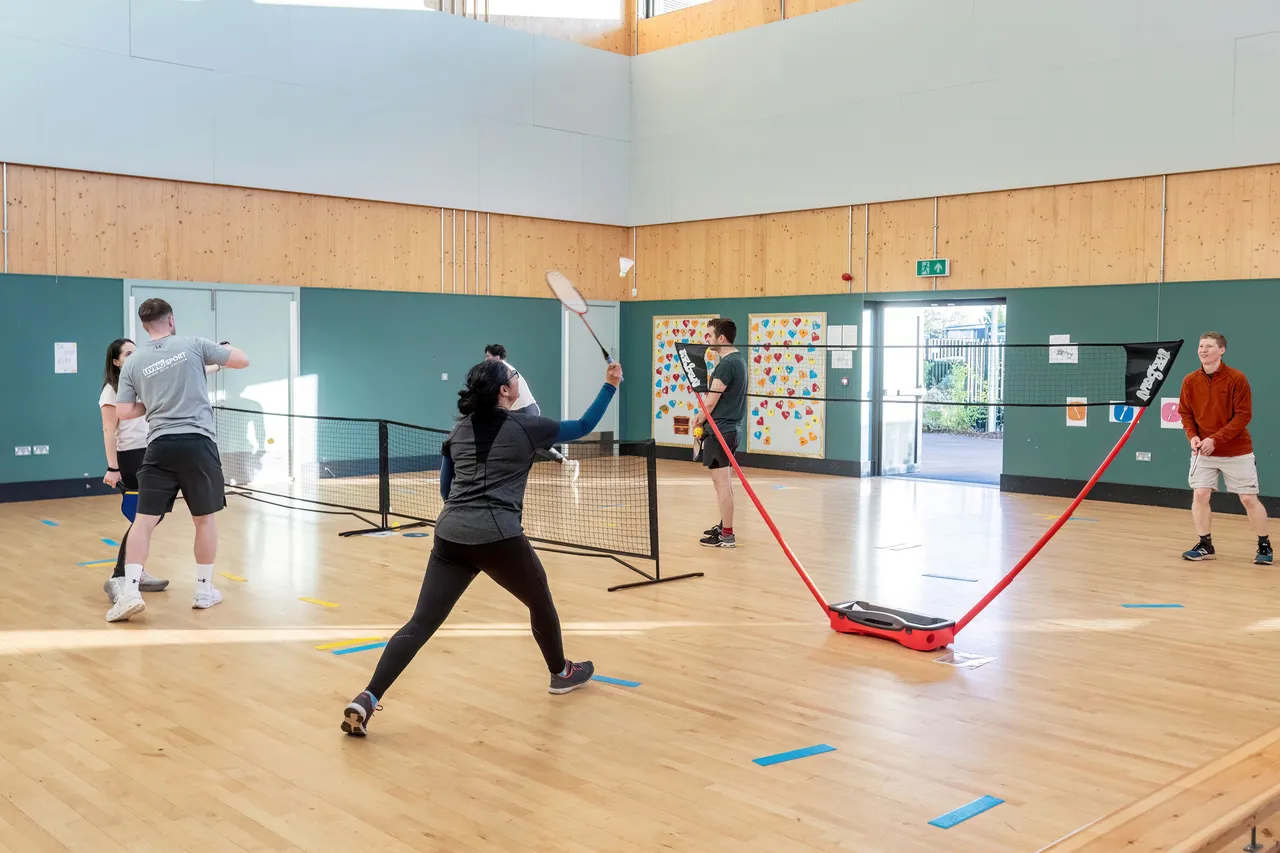 Action shot in a high-ceilinged indoor gym where several people are playing racket sports. In the foreground, a person in dark athletic wear swings a racket toward a shuttlecock. Multiple portable nets are set up across the wooden floor, and other players are visible in various stages of play. The image represents Wintringham residents rally behind racket sports.
