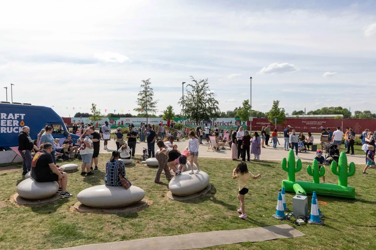An outdoor park setting where Wintringham celebrates the start of summer at Community Day. Families and children gather on a grassy lawn featuring large, smooth, pebble-shaped stone seats. Children play near an inflatable green cactus ring-toss game with blue and white cones, while a crowd of people socialize in the background near food trucks and decorative flags under a lightly clouded sky.