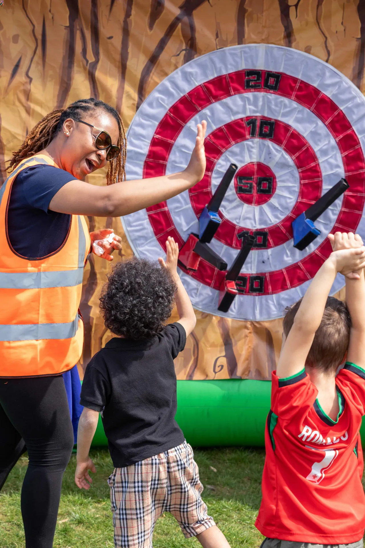 A cheerful scene where Wintringham celebrates the start of summer at Community Day. An event organizer wearing a high-visibility orange vest and sunglasses smiles and gives a high-five to a young child with curly hair. Next to them, another child in a red sports jersey cheers with arms raised in front of a large inflatable target game with velcro axes stuck to it.