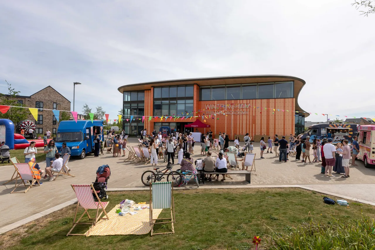 A bustling outdoor festival scene where Wintringham celebrates the start of summer at Community Day. In front of the modern, orange-paneled Wintringham Primary Academy building, a large crowd of families and residents gather on a paved plaza. The event features colorful bunting flags, lounge chairs on a patch of grass in the foreground, and food trucks parked on either side, creating a vibrant community atmosphere under a bright sky.