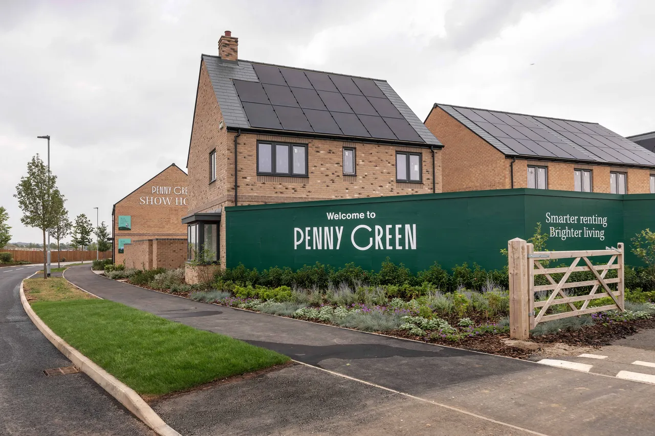An exterior view of a modern housing development entrance featuring a large green hoarding sign that reads "Welcome to Penny Green - Smarter renting, Brighter living". Behind the sign are newly built brick houses fitted with extensive rooftop solar panels, showcasing eco-friendly homes built by the developer