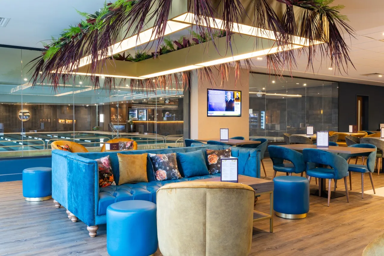 Modern lounge and dining area at David Lloyd St Neots featuring plush blue seating, warm overhead lighting with hanging foliage, and a view of the indoor swimming pool.
