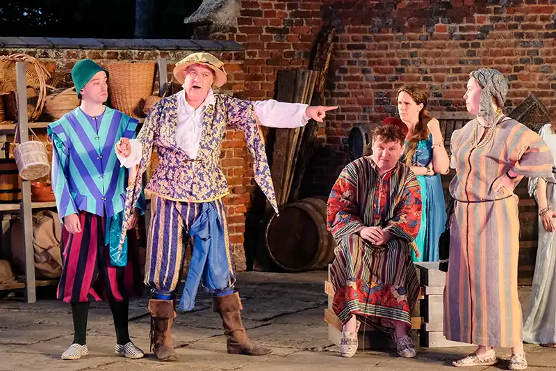 Five actors in colourful Elizabethan-era costumes perform a comedic scene from The Comedy of Errors outdoors at Buckden Towers, with one actor pointing dramatically at another as the others react with surprise and indignation.